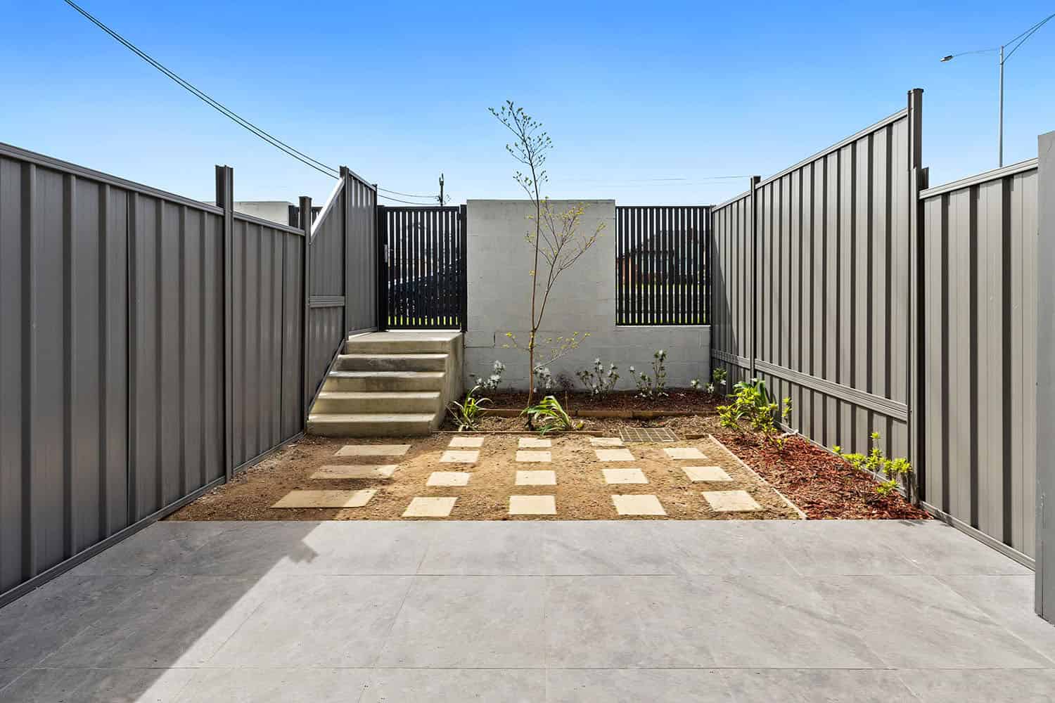 Contemporary backyard featuring a concrete patio, stepping stones, a small tree, and garden beds enclosed by a tall grey metal fence under a clear blue sky.