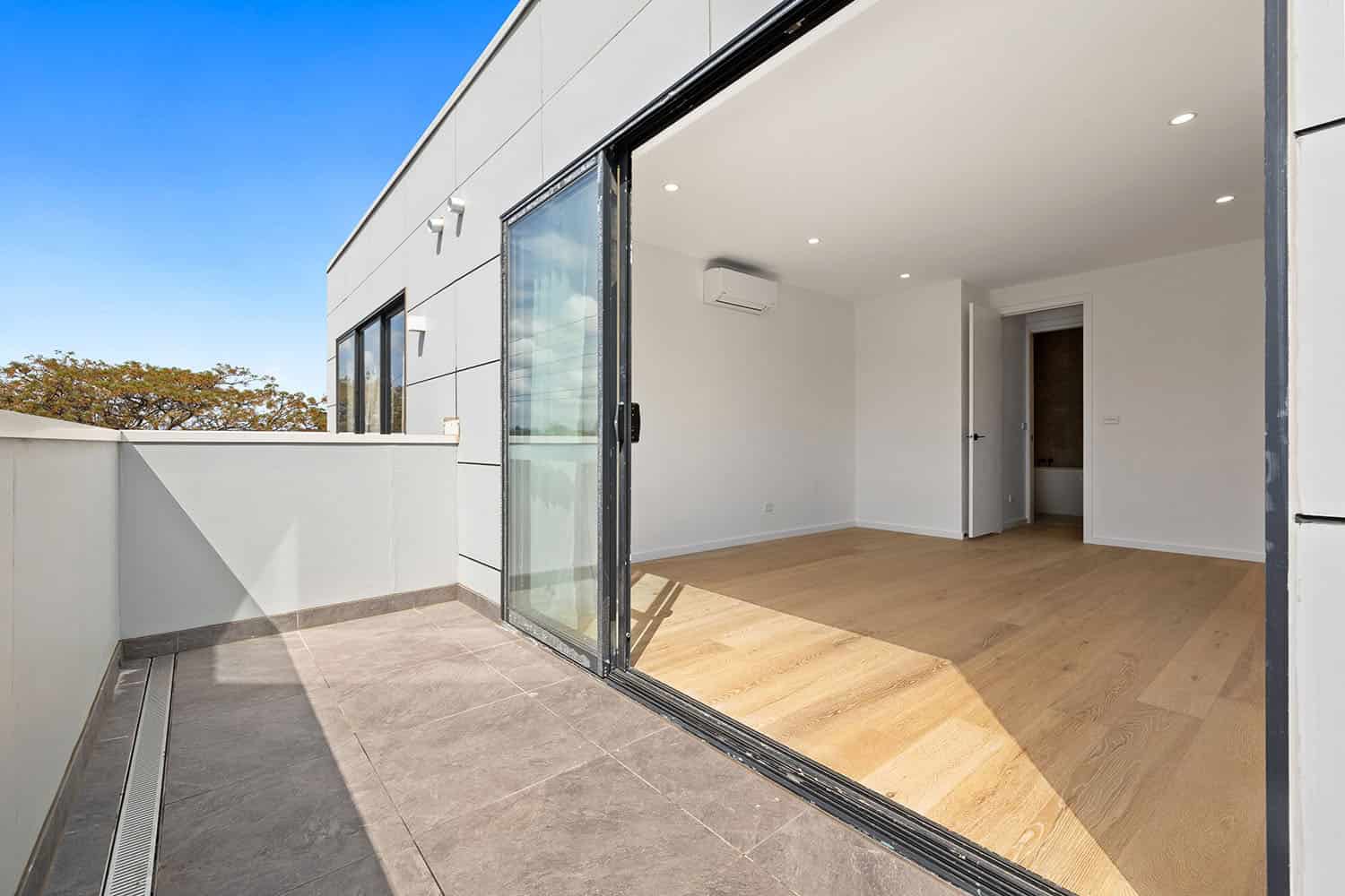 Modern apartment balcony with sliding glass doors leading to an open-plan living area, featuring wooden flooring and white walls, in a contemporary residential property in Ember.