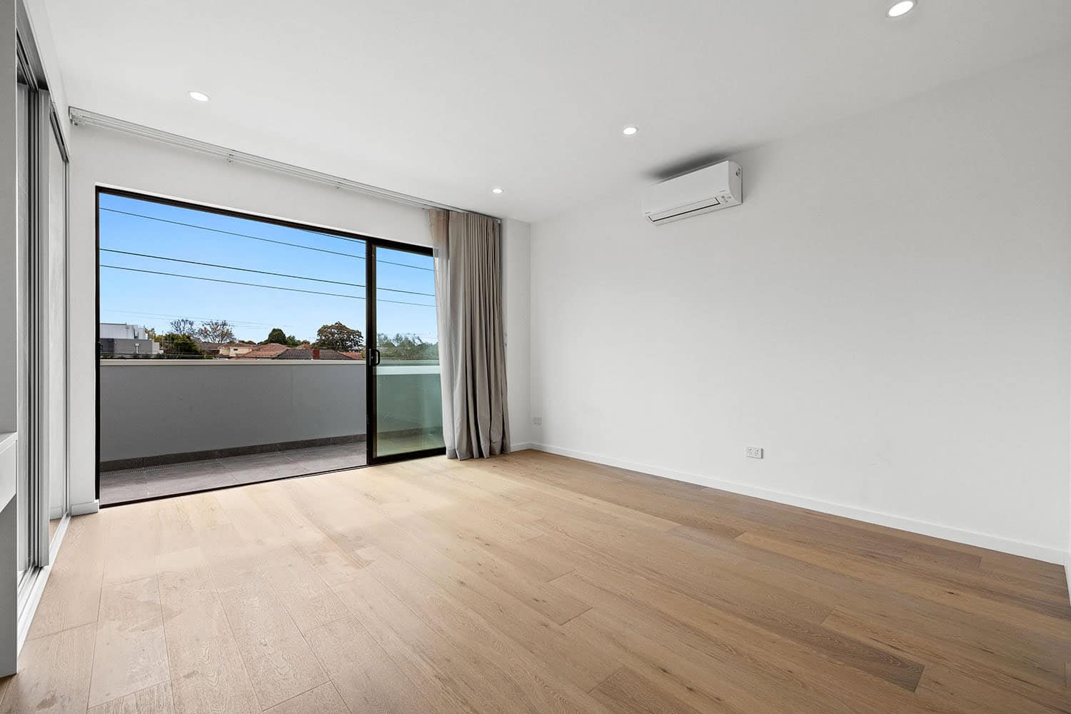 Modern empty living room with large glass sliding doors leading to a balcony, featuring light wooden flooring, white walls, and an air conditioning unit, ideal for property listings.