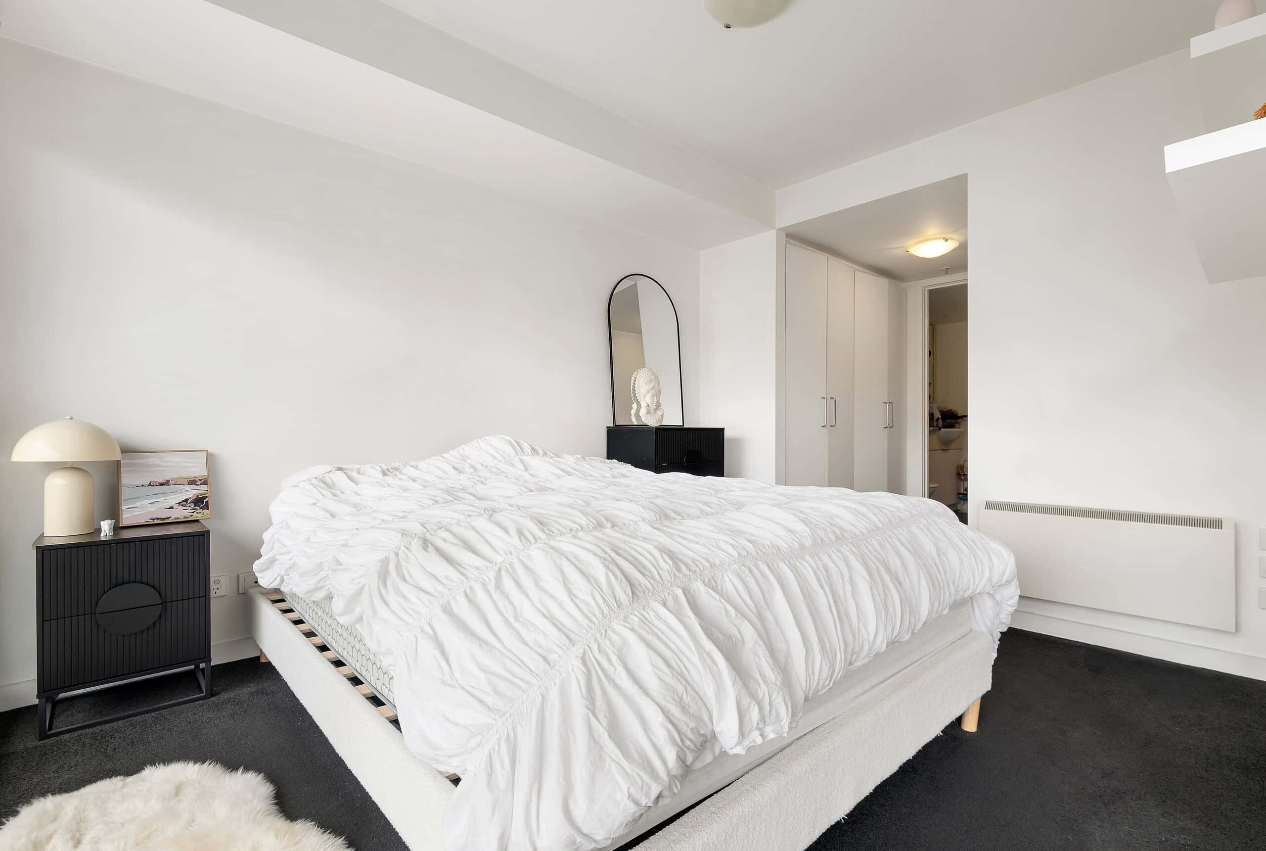 Interior of a modern one bedroom apartment with white walls, a bed with white bedding, and minimal decor.